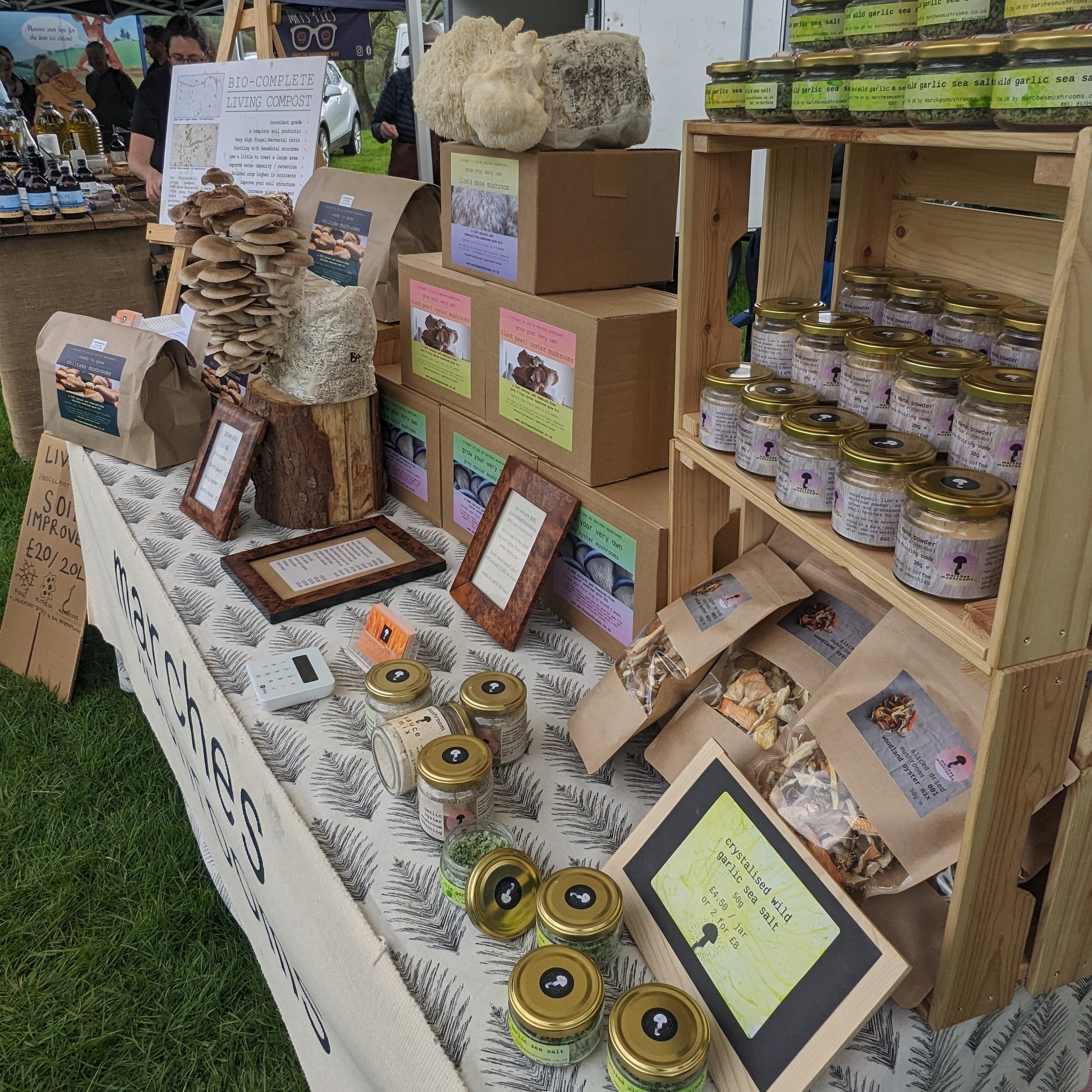 Our gourmet mushrooms growing straight from the hemp substrate block, jars of wild garlic sea salt, seasonings, dried mushrooms and grow-your-own mushroom grow kits, as well as a display board advertising living compost, and informational signs on a table draped with a patterned cloth, set on grass.