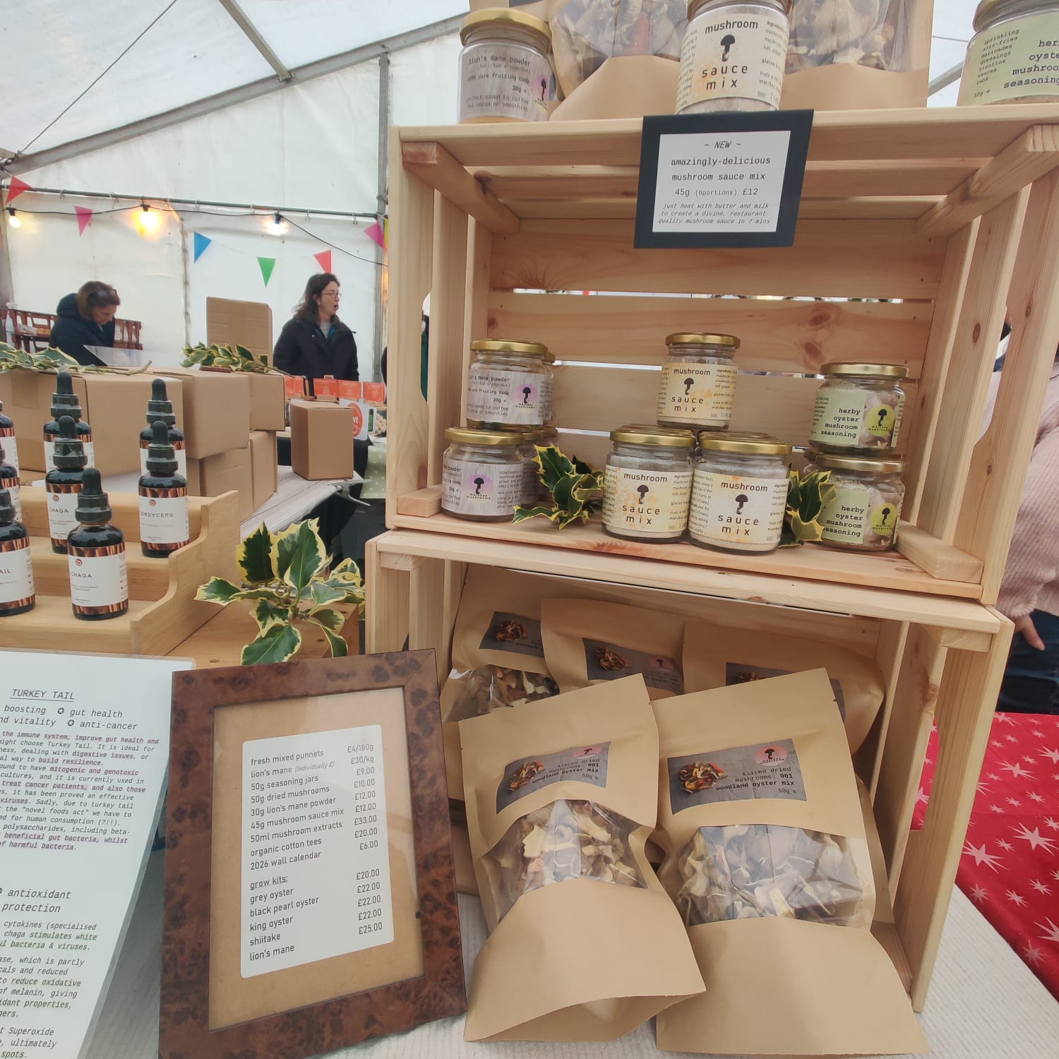 Our high potency extracts and tinctures are available at our market stalls, alongside delicious sauces, seasonings, fresh and dried gourmet mushrooms, growing kits and living compost. A wooden display crate at an outdoor market/christmas fair showcasing a variety of mushroom products from the brand 'marches mushrooms'. The products include jars of sauce mix, tinctures in dark glass bottles, and bags of dried mushrooms in brown paper pouches.
